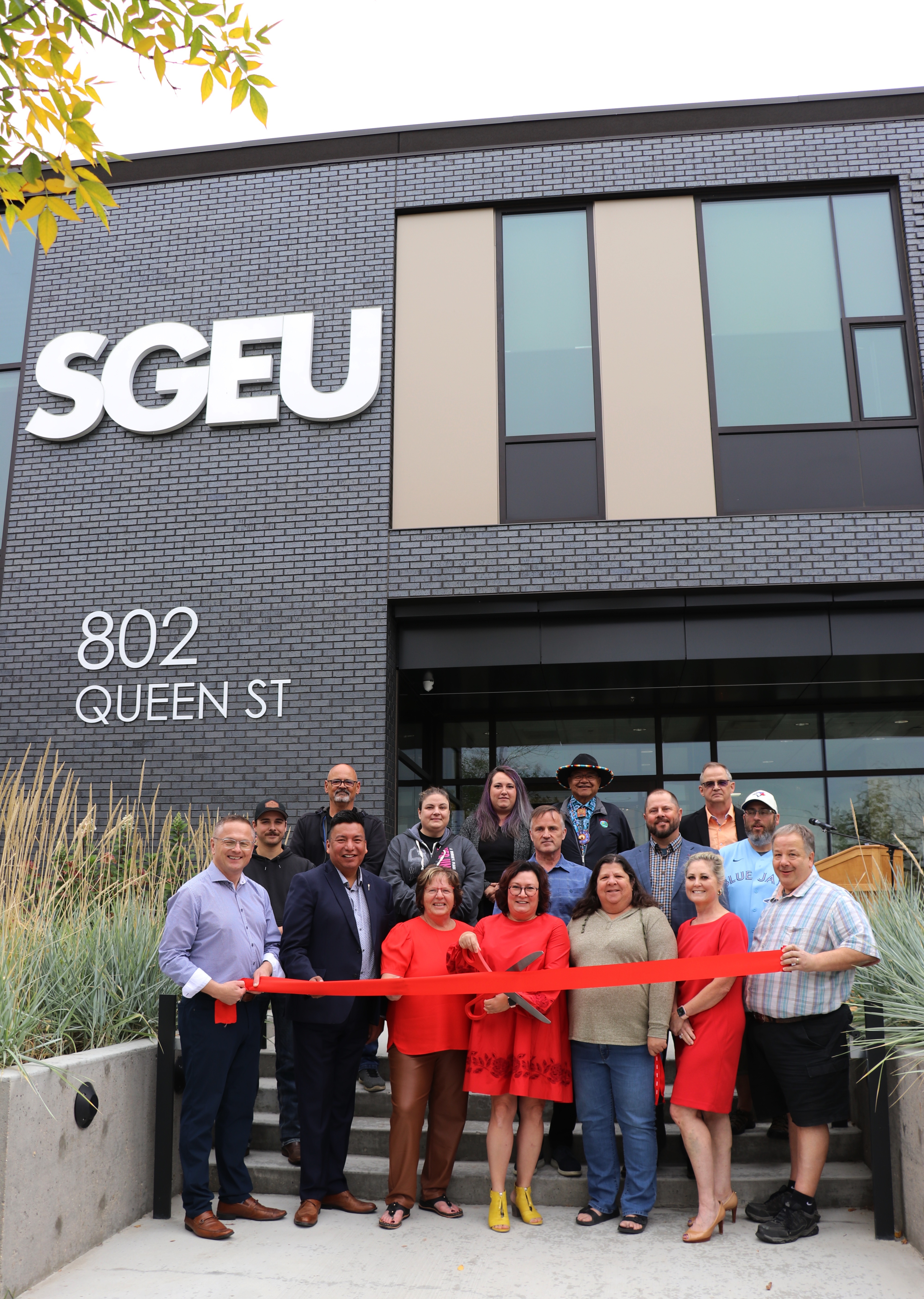 A group of people gathered on the steps of the SGEU front office. There is a red ribbon in front about to be but.